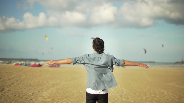 Young sensitive guy looking at kitesurfes flying in the sky under a blue and cloudy sky and opening arms by the sea. Beach in autumn or winter.