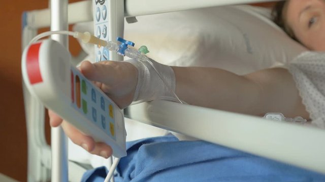 Young woman with drip needle in hand trying to press an emergency button and call for help on a hospital bed. Hospitalized patient with serious injuries feels pain and call a nurse.