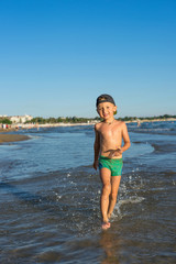 Smiling little baby boy running with splash in the sea on the beach at sunset. Portrait of happy little kid boy on the beach of ocean. Funny cute child making vacations and enjoying summer.