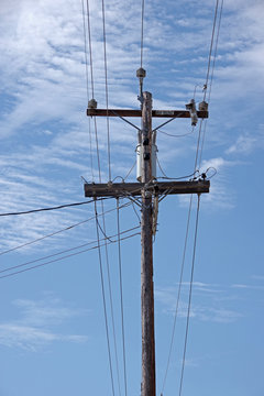 Close view of the top of a tall electricity distribution pole with power lines seen against a blue sky with some white clouds