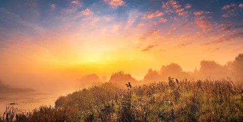 Beautiful golden sunrise panorama at the misty riverside with the bright sun shining through the clouds