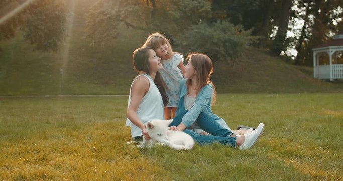 Portrait shot of the cute Caucasian cheerful girls with their pretty mother and haski dog spending time on the green grass in the park in summer and family smiling to the camera.
