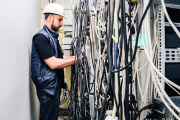 An engineer in a white helmet unravels many wires in a data center. A technician working connects a lot of cables in a server rack. Chaotic plexus electrical wires.