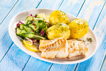 Fried fish with potatoes and vegetable salad on white plate on blue planks