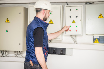 An electrician in a helmet presses the control panel buttons. The specialist works with powerful electric boxes. Engineer checks the electrical supply system.