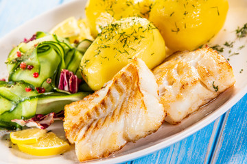 Fried fish with potatoes and vegetable salad on white plate on blue planks