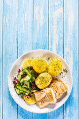 Fried fish with potatoes and vegetable salad on white plate on blue planks