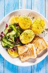 Fried fish with potatoes and vegetable salad on white plate on blue planks