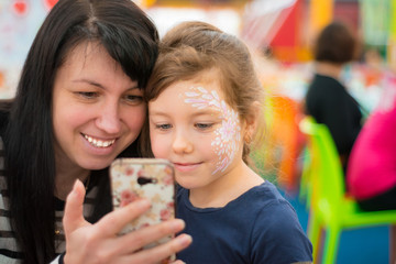 Mom and daughter are looking at a mobile phone. Girls make selfie in a cafe catering. A child in a fashionable makeup with his mother using the phone.