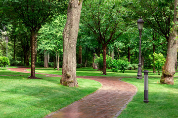 wet after rain winding a pedestrian footpath in a well maintained park with green spaces of green lawn and trees, ground lantern and lamp post, nobody.