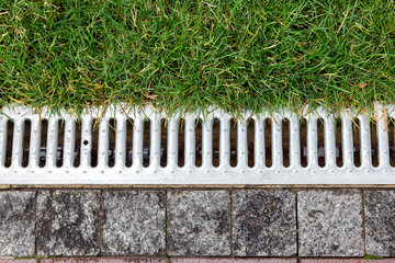 iron grate of a storm drainage system on the side of a footpath made of pavers near a green lawn top view.