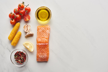 top view of raw salmon steak near lemon, corn, tomatoes, garlic, oil and peppercorns in bowls on marble table