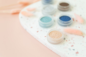 Eyeshadow pots on terrazzo dish, pink background, styled with dried plants, women's makeup flat lay