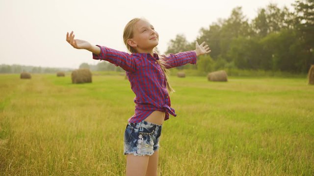 Happy child on field. Adorable cheerful preteen girl in checkered shirt and denim shorts standing with open arms on field with haystacks. Beauty in nature concept