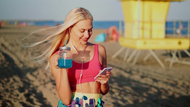 Cheerful young woman drinking refreshing lemonade from a straw at summer by beach side, positive vibes of vacations sunset leisure using smart phone to be always online in social media with earphones