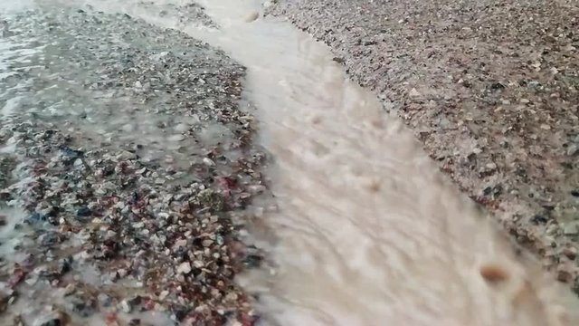 Slow motion muddy water flow through a divet in a concrete driveway or road with rain drops splashing down around
