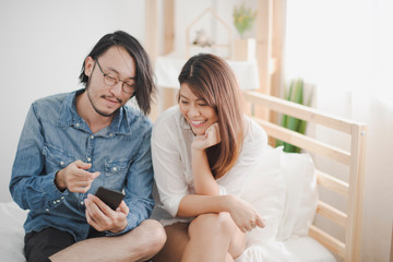 Young couple love Asian man and woman sitting on white bed which looking at smartphone in bed room at home. man and woman glad smiling and felling happy. life of couple love at home.