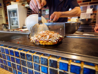 Closeup of a Okonomiyaki cooked on a teppan grill, topped with Katsuobushi fish flakes, sauce, and mayonnaise. Shallow focus. Dotonbori, Osaka, Japan. Travel and cuisine. - 289564639
