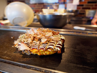 Wide closeup of a fresh-cooked Okonomiyaki topped with Katsuobushi fish flakes, sauce, and mayonnaise, on a Teppan grill. Shallow focus. Dotonbori, Osaka, Japan. Travel and cuisine.
