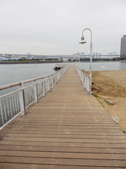 Fototapeta premium Wide vertical view of an old wooden pier with steel railings along the oceanfront bay on a cloudy day. Odaiba Island, Tokyo, Japan