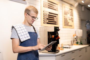 Young barista in workwear using digital tablet to enter new menu of drinks