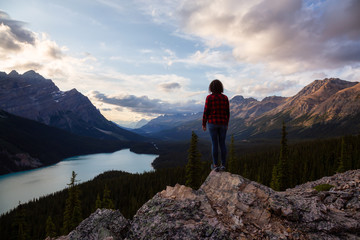 Fototapeta premium Adventurous girl standing on the edge of a cliff overlooking the beautiful Canadian Rockies and Peyto Lake during a vibrant summer sunset. Taken in Banff National Park, Alberta, Canada.