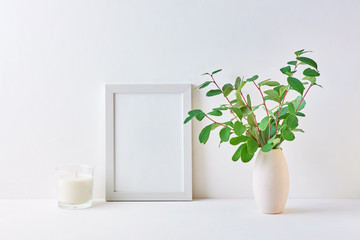 Mockup with a white frame and branches with green leaves in a vase on a white table
