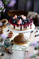 homemade tasty whole cheesecake decorated with figs, blackberries, blueberries and purple sauce on top served on wooden cake stand on grey table with flowers and berries, selective focus