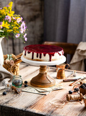 homemade tasty whole cheesecake decorated with purple sauce on top served on wooden cake stand on grey table with flowers and berries, selective focus