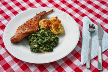 Roasted rabbit leg, marinated in garlic and rosemary served with spinach, chopped onion and potato dumplings on white plate with fork, knife and blue napkin.Ttraditional dish cuisine of Czech republic