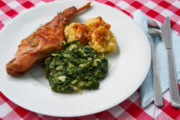 Roasted rabbit leg, marinated in garlic and rosemary served with spinach, chopped onion and potato dumplings on white plate with fork, knife and blue napkin.Ttraditional dish cuisine of Czech republic