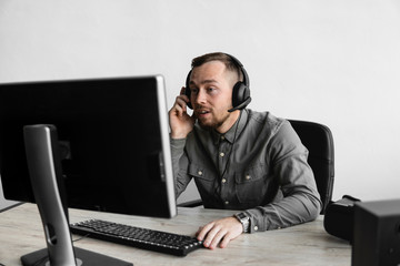 Young businessman or student in a shirt sitting against monitor of computer in headphones and speaking with somebody via internet. Working on a pc at a table in the office.