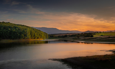 The calm and quiet waters of a mountain lake surrounded by green hills, fields, and forests, distanty mountain range in the background, blue sky with smooth clouds colored in orange by the sunset