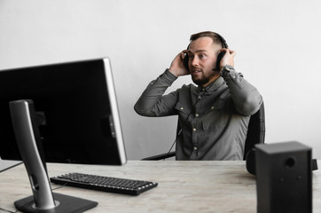 Young businessman or student in a shirt sitting against monitor of computer in headphones and speaking with somebody via internet. Working on a pc at a table in the office.