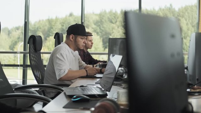 Young man working with laptop looking in monitor. Caucasian male designer in cap using computer for work in office. Medium shot in 4k, UHD
