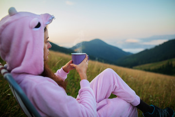 Young girl in a pink dragon costume drinking coffee while sitting in a chair on top of a mountain. Girl in pink pajamas drinks hot drink in the mountains. Incredible mountain landscape at sunrise