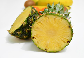 An image of a half-cut pineapple on a white background with bananas, kiwis and mandarins