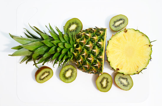 An image of a half-cut pineapple with three half-cut kiwis nearby on a white breadboard on a white background