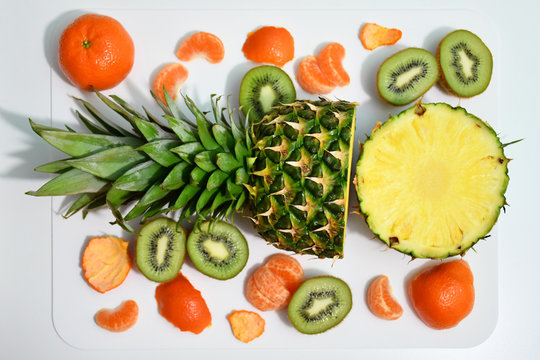 An image of a half-cut pineapple, three half-cut kiwis, one pineapple, a few segments of tangerines and a few tangerine rinds nearby on a white breadboard on a white background