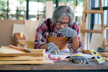 Old Asian craftsman touch his mouth and thinking about his process for working with wood product in the room during day time.