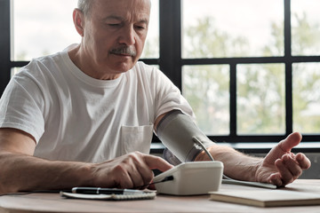 Old hispanic man feeling bad using a home blood pressure machine to check his health in the morning