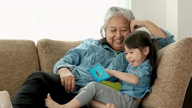 Asian grandmother or old woman sit on sofa and enjoy with her little child with image book for kids and they look fun and happy together.