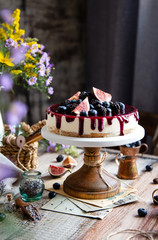 homemade tasty whole cheesecake decorated with figs, blackberries, blueberries and purple sauce on top served on wooden cake stand on grey table with flowers and berries, selective focus