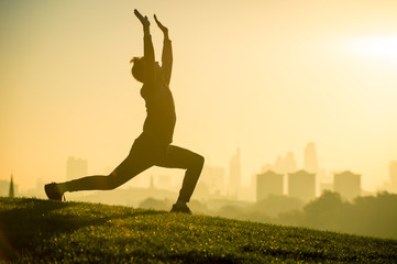 Scenic sunrise silhouette of a man stretching in a relaxed Anjaneyasana yoga lunge on the grassy top of a hill front of a misty golden view of the London city skyline