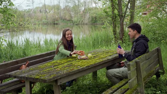 Girl and guy have picknic with apples, benzel and hot tea in thermos. They sitting on old wooden benchs and table which are covered with green moss