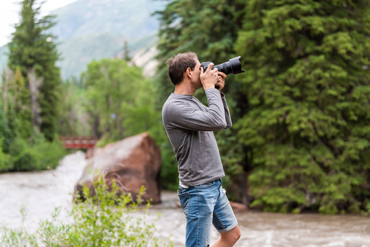 Man taking photos with camera in Redstone, Colorado during summer with large boulder and red bridge on Crystal river by trees