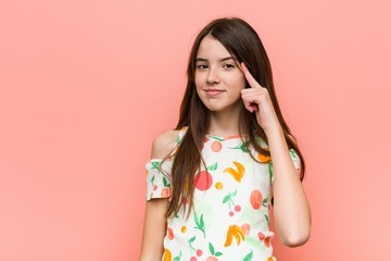 Girl wearing a summer clothes against a red wall pointing temple with finger, thinking, focused on a task.