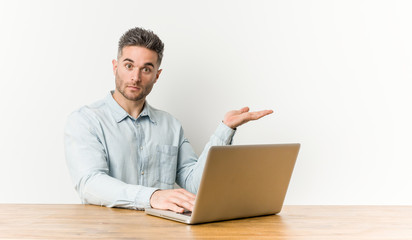 Young handsome man working with his laptop showing a copy space on a palm and holding another hand on waist.