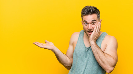 Young fitness man against a yellow background holds copy space on a palm, keep hand over cheek. Amazed and delighted.