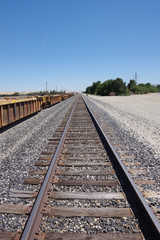 Fototapeta premium A landscape scene looking south with railroad tracks lined by a chain of seemingly forgotten rail transport wagons; mountains and trees in the central California inland distance and blue sky above